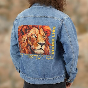 Op de afbeelding: Lichtblauwe spijkerjas met een gedetailleerde illustratie van een leeuwenkop in oranje en bruine tinten. De tekst "RISING LION" en Hebreeuwse tekst zijn in geel gedrukt.