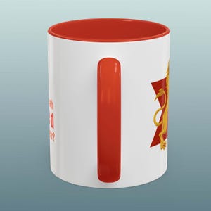 May include: White ceramic mug with a red interior and handle. The mug features a gold lion design on a red starburst shape. Text on the side of the mug reads, "What's with the red star?"