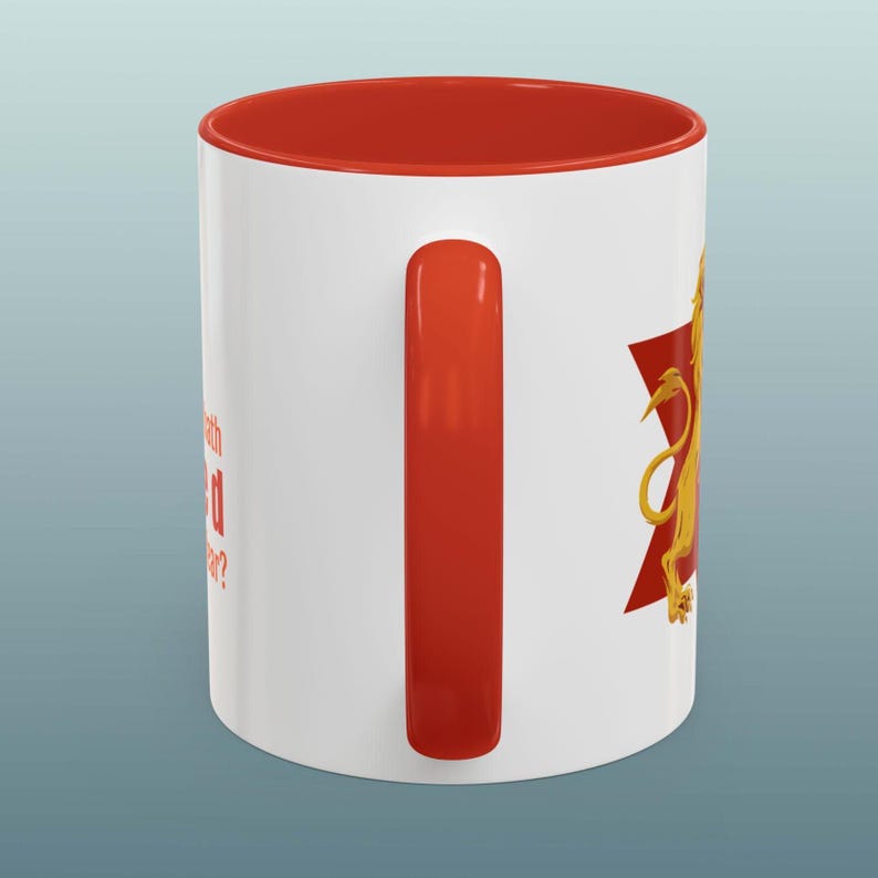 May include: White ceramic mug with a red interior and handle. The mug features a gold lion design on a red starburst shape. Text on the side of the mug reads, "What's with the red star?"