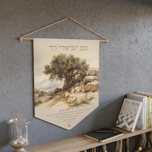 May include: A beige banner with a landscape painting of a tree and rocks, with text in Hebrew and English. The banner is hung from a wooden rod with a string. The text includes "AMPS 9:15" and a quote. The banner is displayed on a gray wall.