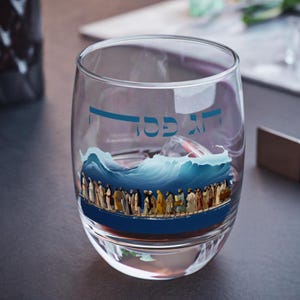 Könnte beinhalten: Ein klares Glas mit einem blauen und weißen Bild einer Welle und Menschen, die auf dem Wasser gehen. Der Text "אך כסה" ist auf das Glas gedruckt.