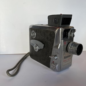 May include: A silver Keystone Bel Air 8 mm movie camera with a black leatherette body and a built-in flash. The camera has a lens with a zoom ring and a strap attached.