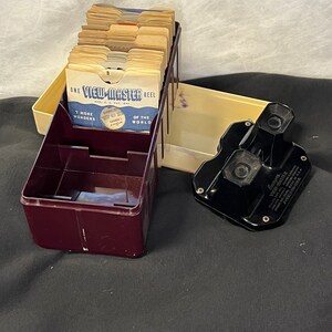 May include: A vintage View-Master reel viewer with a burgundy plastic case and a black plastic viewer. The case is filled with a stack of View-Master reels. The reels have images of "7 More Wonders of the World".