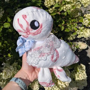 May include: A plush toy in the shape of a creature with a white body and pink accents. It has a large black eye and a blue and white checkered bow. The toy is decorated with lace and floral patterns.