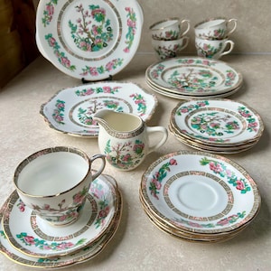 Könnte beinhalten: Vintage Porzellan-Teeservice mit floralem Design in Rosa und Grün. Das Set umfasst Tassen, Untertassen, Teller und einen Sahnekännchen, alle mit Goldrand. Die Teller haben einen gewellten Rand.