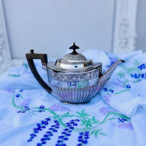 May include: A silver teapot with an ornate design sits on a blue and white floral tablecloth. The teapot has a rounded body and a spout on the right side. The handle is made of dark wood.