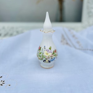 May include: A white porcelain bottle with a floral design and gold accents. The bottle has a pointed top and a wide base. It features a floral pattern with pink, yellow, and blue flowers. The bottle has a gold rim and a gold logo on the bottom.