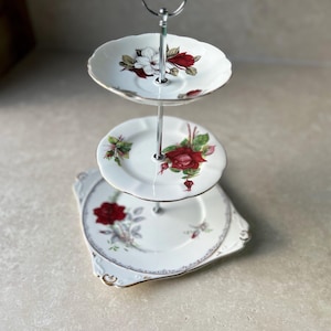 Può includere: Un alzatina a tre livelli con motivi floreali. I piatti sono bianchi con rose rosse e bordi dorati. Il supporto ha una maniglia e un supporto in metallo argentato.