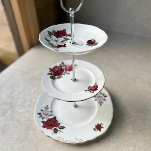 Può includere: Un alzatina a tre livelli con piatti in porcellana bianca. Ogni piatto è decorato con rose rosse e bordi dorati. L'alzatina ha un manico e una struttura di supporto in metallo argentato.