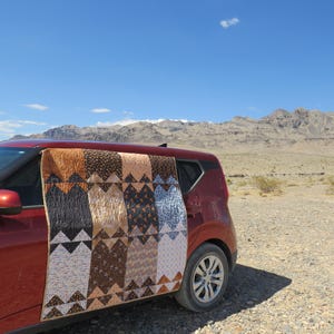 May include: A rectangular quilt featuring a repeating geometric pattern of triangles in shades of brown, tan, black, and white. The quilt is draped over the side of a red car, displaying a multi-colored patchwork design with various small prints.
