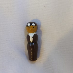 May include: A wooden figurine of a person wearing a brown leather jacket and goggles. The figurine has a white face with black eyes and a brown mouth.