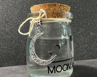 Könnte beinhalten: Ein klares Glas mit einem Korken, gefüllt mit Flüssigkeit. Ein silberner Halbmond-Anhänger hängt an dem Glas. Das Glas hat eine schwarze Grafik eines Halbmonds und das Wort "MOON".