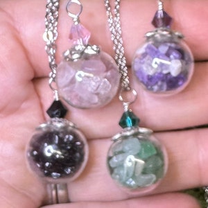 Puede incluir: Cuatro collares con globos de cristal llenos de piedras preciosas de colores. Cada globo cuelga de una cadena plateada y tiene una tapa plateada decorativa y una cuenta de cristal. Las piedras son rosas, moradas, negras y verdes.