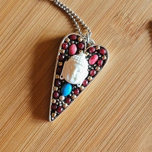 May include: A silver chain necklace with a heart-shaped pendant. The pendant is decorated with red, pink, and blue gemstones and features a white Buddha head in the center.