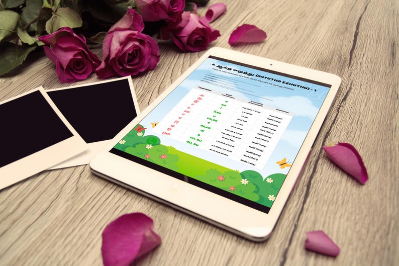 May include: A white tablet displaying a colorful educational chart with text in a foreign language. The tablet is surrounded by pink roses, photo frames, and rose petals on a wooden surface.