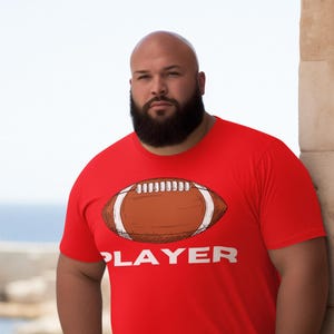 May include: Red t-shirt with a football graphic and the word "PLAYER" in white. The shirt is short-sleeved and worn by a person. The background includes a stone structure and a body of water.