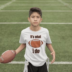May include: A person wearing a white t-shirt with the text "it's what I do" and a football graphic. The person is holding a brown football on a green field.