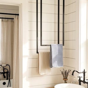 May include: A black metal ceiling-mounted towel rack with a rectangular frame design. Two towels are hanging from the bottom bar, one with thin beige stripes and the other with bold navy blue and white horizontal stripes, positioned above a wooden vanity.