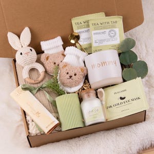 May include: A gift box filled with baby items and self-care products. Includes a crochet bunny rattle, bear-faced baby booties, a mug with "mommy" written on it, tea packets, a candle, and eye masks. The box is on a white, fluffy surface.