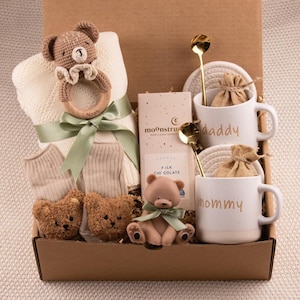 May include: A baby gift box containing a cream blanket, socks, and teddy bear toys. The box also includes two white mugs with "daddy" and "mommy" printed on them, chocolate, and gold spoons. A wooden baby rattle is also included in the gift set.