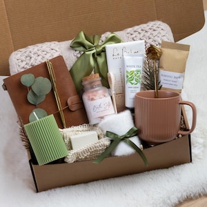 K&ouml;nnte beinhalten: Eine Geschenkbox mit Pflegeprodukten. Sie enth&auml;lt ein braunes Lederjournal, eine gr&uuml;ne Kerze, Badesalz, Handcreme, einen Becher und ein kleines Handtuch. Die Box ist mit einem gr&uuml;nen Band gebunden und enth&auml;lt eine wei&szlig;e Decke.