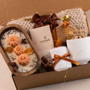 May include: A gift box containing a wooden dough bowl candle with three pumpkin-shaped wax inserts, a Moonstruck chocolate bar, a white ribbed ceramic mug, a white fuzzy cloth with a brown ribbon, a beige knit blanket, and a gold spoon.