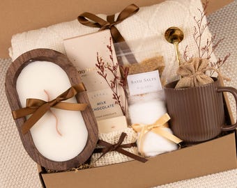 May include: A gift box containing a cream-colored knit blanket, a dark brown ribbed mug with a gold spoon, a wooden oval candle, a milk chocolate bar, a small bag of bath salts, and a small burlap pouch, all tied with brown satin ribbons.
