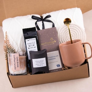 May include: A gift box filled with a white knit blanket, coffee, chocolate, a candle, and a mug. The mug is peach-colored with vertical ridges. A gold spoon rests inside the mug, and a small coaster sits beside it.
