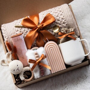 May include: A gift box filled with a variety of items, including a knitted blanket, a candle, a mug, a chocolate bar, a small stuffed bunny, and a wooden rattle. The box is decorated with a brown ribbon.