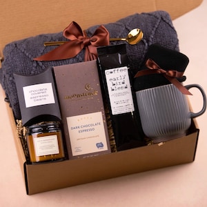 May include: A gift box filled with a gray knit blanket, a gray mug, a jar of chocolate-covered espresso beans, a bar of dark chocolate espresso, and a bag of coffee. A gold spoon and a brown ribbon are included.