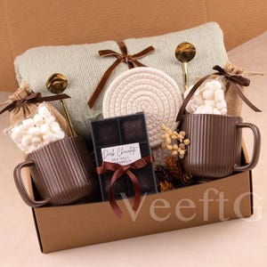 Puede incluir: Una caja de regalo con tem&aacute;tica acogedora. Incluye dos tazas marrones acanaladas, malvaviscos, chocolate negro, una manta verde claro y un posavasos tejido. Cucharas doradas y detalles decorativos completan el conjunto.
