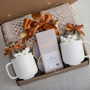 Puede incluir: Una caja de regalo con dos tazas blancas acanaladas llenas de malvaviscos, cucharas doradas y una barra de chocolate con leche "Moonstruck". Una manta de punto trenzado beige y un lazo naranja completan la presentaci&oacute;n. Las luces de hadas a&ntilde;aden un toque festivo.