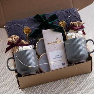 May include: A gift box filled with a gray knit blanket, two gray mugs, marshmallows, chocolate, and gold spoons. The box is tied with a green velvet bow and contains decorative string lights.