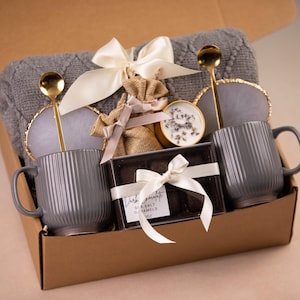 May include: A gift box filled with a gray knitted blanket, two gray ribbed mugs, gold spoons, coasters, a candle, and a box of dark chocolate caramels. The box is tied with a white ribbon.