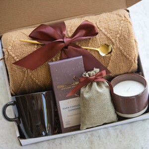Peut inclure: Une bo&icirc;te-cadeau remplie d'articles confortables pour prendre soin de soi, comprenant une couverture en tricot marron, une tasse en c&eacute;ramique marron, une barre de chocolat noir expresso, un petit sac en toile de jute avec un ruban marron et une bougie en c&eacute;ramique marron.