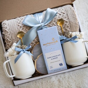 May include: A gift box with a cream-colored cable-knit blanket, two white ribbed mugs with marshmallows, gold spoons, a bar of dark chocolate, and a blue ribbon. The chocolate bar reads "Moonstruck Portland Design" and "Dark Chocolate Almond".