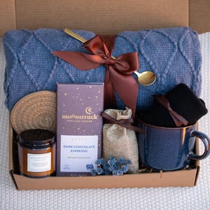 May include: A gift box filled with a blue knit blanket, a dark chocolate bar, a candle, a mug, a small bag of coffee beans, a coaster, and a gold spoon. The box is tied with a brown ribbon.