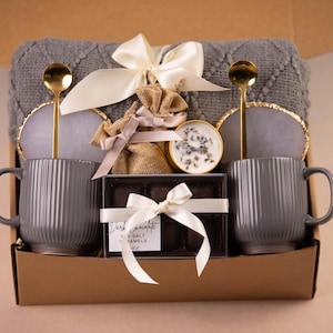 May include: A gift box with two gray ribbed mugs, gold spoons, coasters, and a gray blanket. The box also contains a small candle, a burlap bag, and a box of dark chocolate sea salt caramels, all tied with white ribbons.