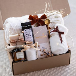 May include: A gift box containing a white textured throw blanket with a rust-colored velvet bow, a bag of Early Bird Blend coffee, a Moonstruck milk chocolate bar, a white ribbed mug, a Rich Mahogany candle, and a small jar with a wooden spoon.