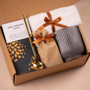 Peut inclure: Une bo&icirc;te cadeau en carton marron contenant une tasse en c&eacute;ramique grise, un petit sac en toile de jute avec un ruban marron, une cuill&egrave;re dor&eacute;e et un paquet de m&eacute;lange de chai caramel latte. La bo&icirc;te est ouverte et les articles sont visibles &agrave; l'int&eacute;rieur.