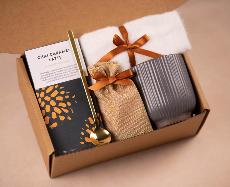 Peut inclure: Une bo&icirc;te cadeau en carton marron contenant une tasse en c&eacute;ramique grise, un petit sac en toile de jute avec un ruban marron, une cuill&egrave;re dor&eacute;e et un paquet de m&eacute;lange de chai caramel latte. La bo&icirc;te est ouverte et les articles sont visibles &agrave; l'int&eacute;rieur.