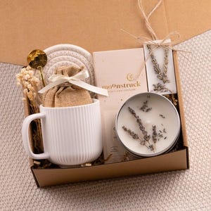 May include: A gift box filled with a white mug, a small burlap bag tied with a ribbon, a gold spoon, a candle, a coaster, and a card. The box is brown and contains dried flowers and lavender sprigs. The mug has vertical lines.