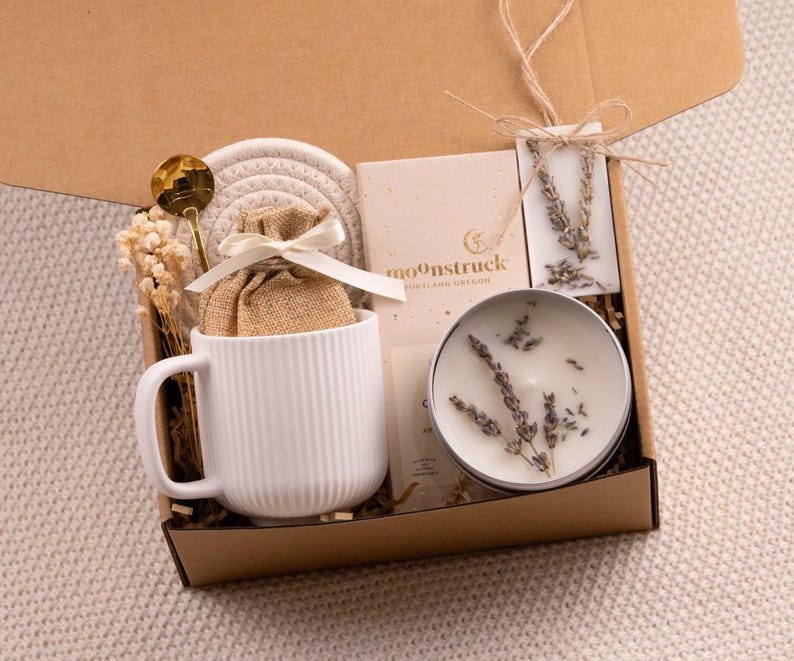 May include: A gift box filled with a white mug, a small burlap bag tied with a ribbon, a gold spoon, a candle, a coaster, and a card. The box is brown and contains dried flowers and lavender sprigs. The mug has vertical lines.