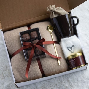 Peut inclure: Une bo&icirc;te-cadeau remplie d'une couverture en laine brune, une tasse, un petit sac de chocolats, une bougie et une cuill&egrave;re en or. La bo&icirc;te est attach&eacute;e avec un ruban brun et un n&oelig;ud.