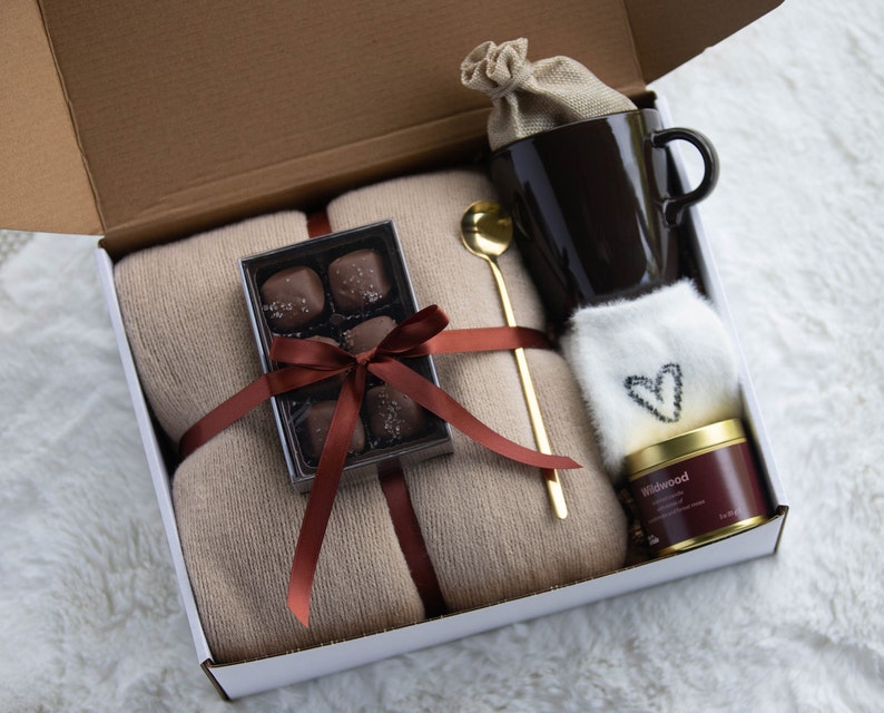 Peut inclure: Une bo&icirc;te-cadeau remplie d'une couverture en laine brune, une tasse, un petit sac de chocolats, une bougie et une cuill&egrave;re en or. La bo&icirc;te est attach&eacute;e avec un ruban brun et un n&oelig;ud.