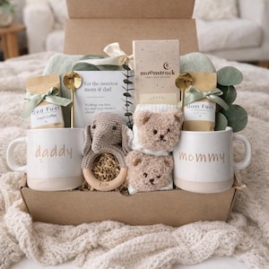 May include: A gift box containing two white ceramic mugs labeled daddy and mommy, two bags of coffee labeled Dad Fuel and Mom Fuel, a Moonstruck chocolate bar, a crochet elephant rattle, two fuzzy bear-shaped socks, and two gold spoons.