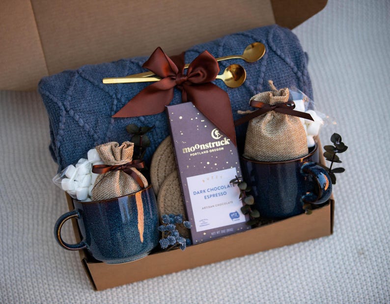 May include: A gift box filled with a blue cable-knit blanket, two blue ceramic mugs, a bar of dark chocolate, two gold spoons, and two small burlap bags filled with marshmallows. The box is tied with a brown satin ribbon.