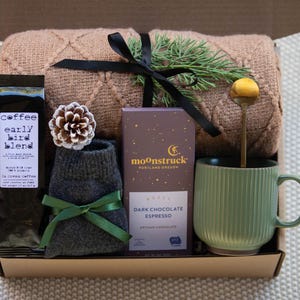 Peut inclure: Une bo&icirc;te-cadeau remplie d'articles confortables pour une soir&eacute;e relaxante. Elle comprend une couverture en tricot marron, une paire de chaussettes grises, un sac de caf&eacute; &eacute;tiquet&eacute; "coffee early bird blend", une barre de chocolat noir espresso artisan, une tasse en c&eacute;ramique verte et une cuill&egrave;re dor&eacute;e.