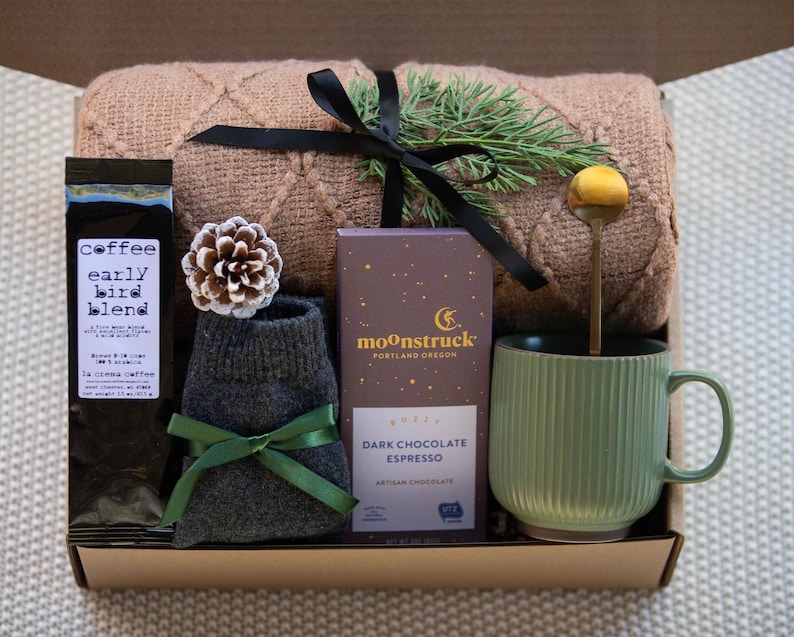 Peut inclure: Une bo&icirc;te-cadeau remplie d'articles confortables pour une soir&eacute;e relaxante. Elle comprend une couverture en tricot marron, une paire de chaussettes grises, un sac de caf&eacute; &eacute;tiquet&eacute; "coffee early bird blend", une barre de chocolat noir espresso artisan, une tasse en c&eacute;ramique verte et une cuill&egrave;re dor&eacute;e.