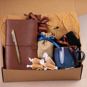 Peut inclure: Une bo&icirc;te en carton marron remplie de divers cadeaux, dont un journal en cuir marron avec un stylo dor&eacute;, une tasse en c&eacute;ramique bleue, un petit sac en toile de jute avec un ruban dor&eacute;, des bonbons au caramel et une couverture en tricot marron.